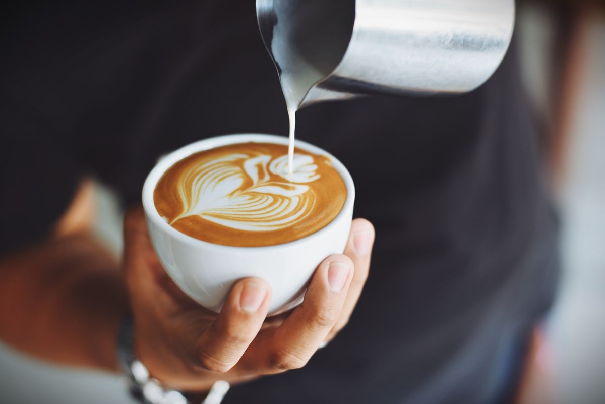 Hand pouring steamed milk into a white cup to finish latte art on coffee